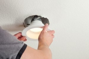 A person is installing a high-efficiency LED recessed light fixture in a ceiling. The focus is on the hands as they secure the light into place, showcasing a typical home electrical maintenance scenario. electrical repair, electrical residential, LED light installation, recessed lighting, home improvement, energy efficiency, Sacramento electrician, Roseville electrical service, Rocklin lighting solutions, Folsom home maintenance, Auburn electrical contractor, Citrus Heights lighting installation